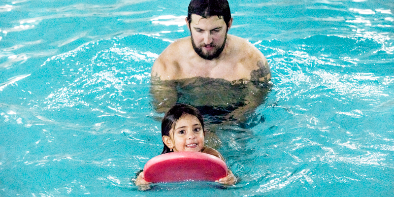 A smiling girl kicks with a pink kickboard while her coach swims behind her