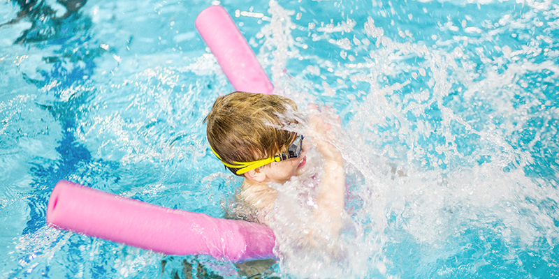 A child splashes through the water with a pink pool noodle