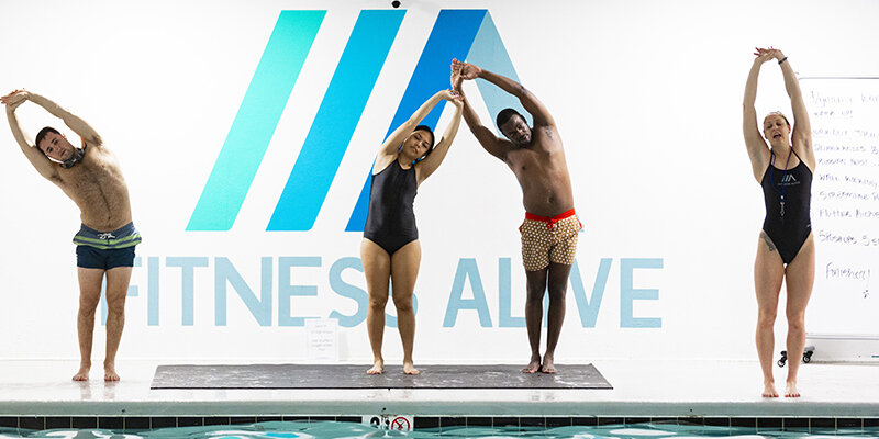 Adult swimmers stretching on the pool deck in front of the Fitness Alive logo