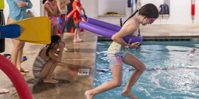 A camper jumps into the pool while others wait on the deck with kickboards