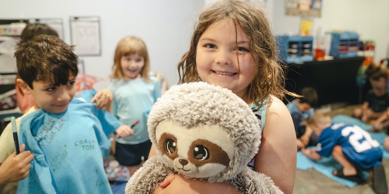 A smiling camper holds up a stuffed sloth indoors