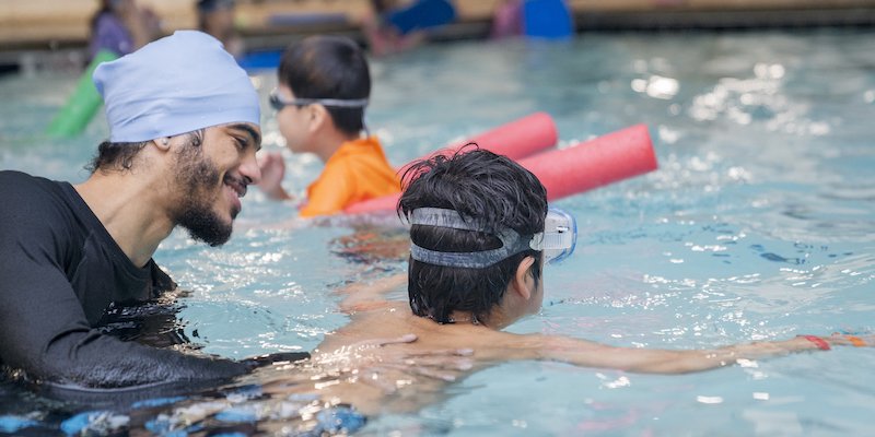 A smiling swim coach supports a child practicing a front float during a lesson